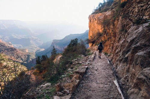 A hiker walks along a narrow trail beside a rocky cliff, with a vast canyon view in the background.