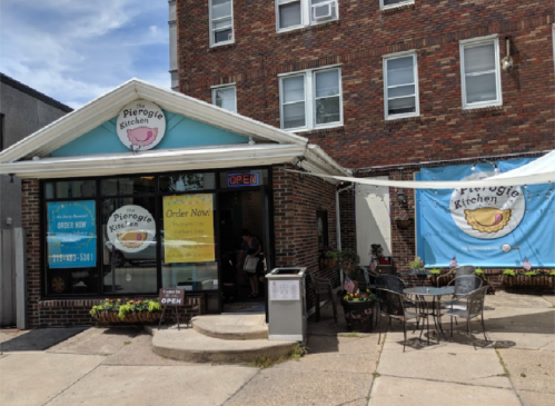 Exterior of Pierogi Kitchen with a blue awning, open sign, and outdoor seating area. Bright and inviting atmosphere.