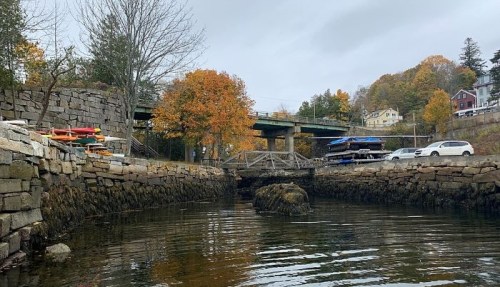 A calm waterway with autumn trees, a small bridge, and parked boats under a cloudy sky.