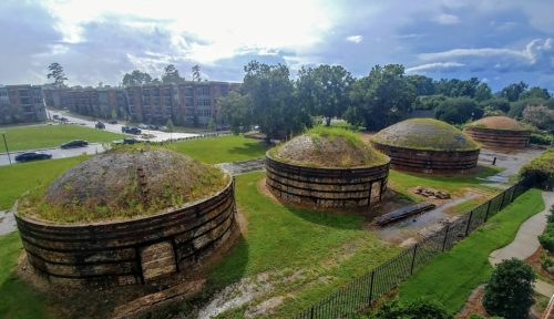 Abandoned circular structures with grassy tops, surrounded by greenery and a residential area in the background.