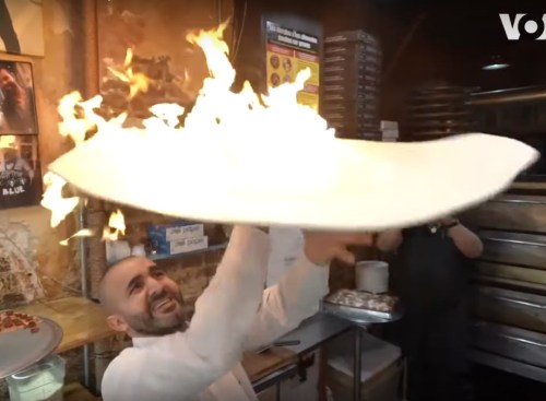 A chef joyfully tosses a flaming pizza in a bustling kitchen, showcasing a dramatic cooking technique.