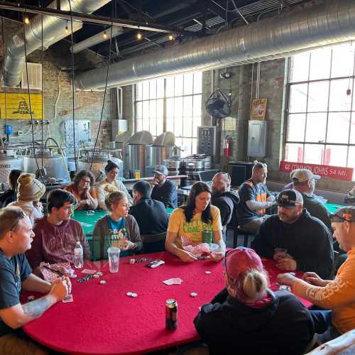 A group of people playing poker around a red table in a spacious, industrial-style room with large windows.