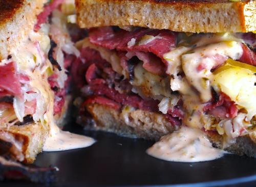 Close-up of a stacked sandwich with layers of pastrami, sauerkraut, and creamy sauce between toasted bread.