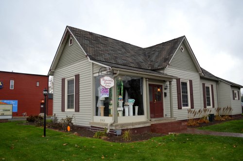 A quaint, light-colored house with a sign reading "Handcrafted Gifts" in the window, surrounded by green grass.