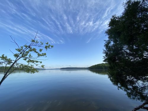 A serene lake scene with calm water, blue sky, and wispy clouds, framed by trees on either side.