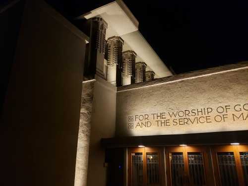 A well-lit building at night featuring architectural details and a sign that reads, "For the worship of God and the service of man."