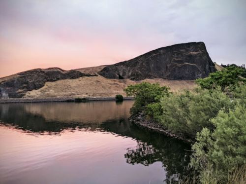A serene landscape featuring a calm lake reflecting a rocky hill and soft pink sky, surrounded by greenery.