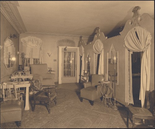 A vintage living room with ornate curtains, chairs, and a small table, featuring a warm, inviting atmosphere.