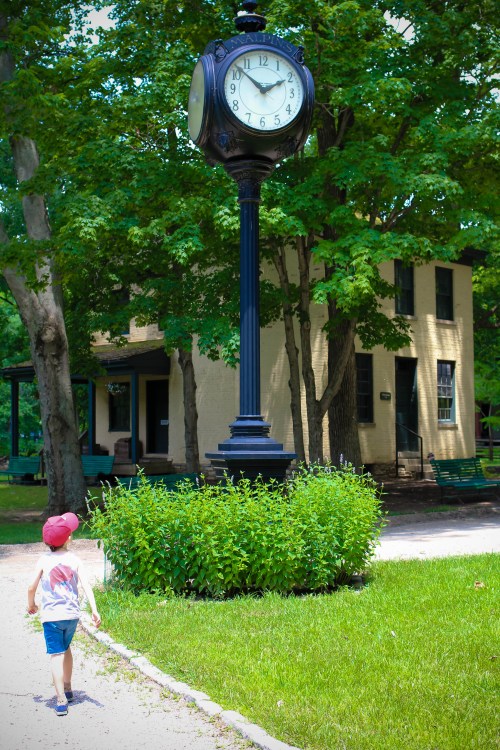 A child in a cap walks towards a large clock surrounded by greenery, with a building visible in the background.