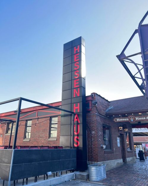 Sign for Hessen Haus, a brick building with outdoor seating, under a clear blue sky.