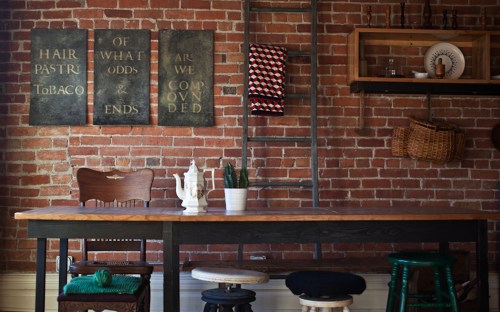 A rustic dining area with a wooden table, mixed seating, and vintage wall art against a brick backdrop.