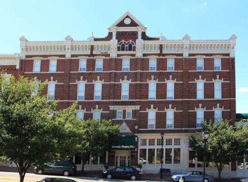 Historic brick hotel with ornate architectural details, surrounded by trees and parked cars under a clear blue sky.