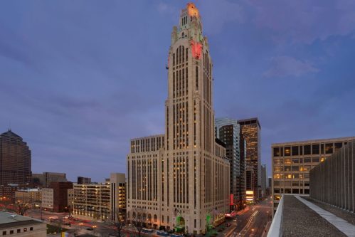 A tall, illuminated skyscraper at dusk, surrounded by modern buildings and city streets.