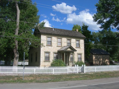 A two-story stone house with a front porch, surrounded by trees and a white picket fence, under a blue sky.