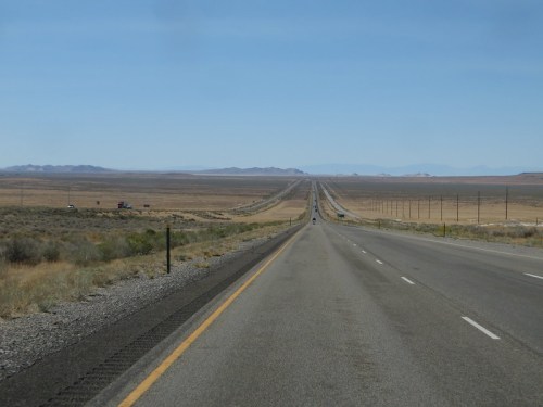 A long, straight highway stretches through a vast, arid landscape under a clear blue sky.