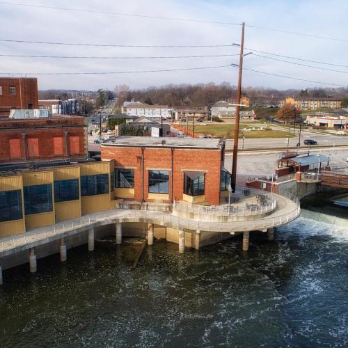 Aerial view of a riverside building with a curved deck, surrounded by water and a small town in the background.
