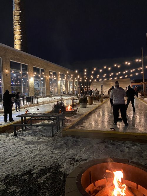 A winter scene with people enjoying an outdoor gathering by a fire pit, string lights, and an ice rink at night.