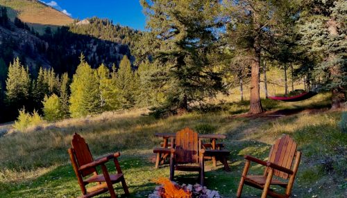 A serene campsite with wooden chairs around a fire pit, surrounded by trees and mountains under a clear blue sky.