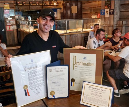 A man smiles while holding framed proclamations and citations in a café, with people dining in the background.