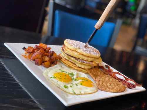 A plate of breakfast featuring pancakes, eggs, bacon, hash browns, and seasoned potatoes, with a knife in the pancakes.