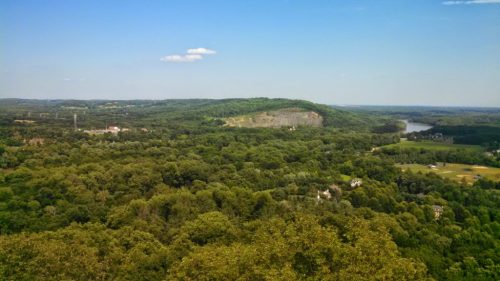 A panoramic view of lush green hills and a river under a clear blue sky, with scattered clouds in the distance.