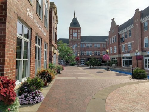 A charming brick pathway lined with flowers, leading to a clock tower and buildings in a quaint town square.