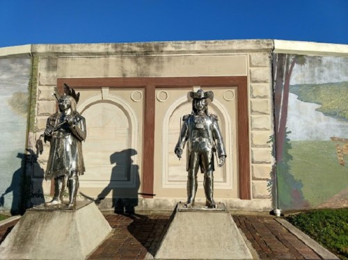 Two metallic statues stand side by side against a mural, one resembling a Native American and the other a colonial figure.