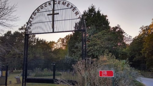 Entrance gate of a cemetery with a cross, surrounded by trees and a sign reading "318 Rotary Ln." at sunset.