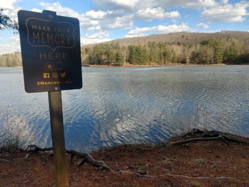 A sign by a lake encourages visitors to make memories, with trees and hills in the background under a blue sky.