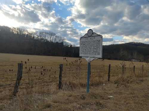 Historical marker in a field with a cloudy sky and mountains in the background. Fenced area with dry grass.