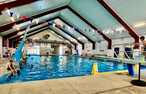 Indoor swimming pool with colorful bunting, diving board, and people swimming and relaxing around the water.