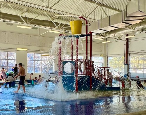 Indoor water play area with a large splash feature, children playing, and a man standing nearby. Bright and lively atmosphere.