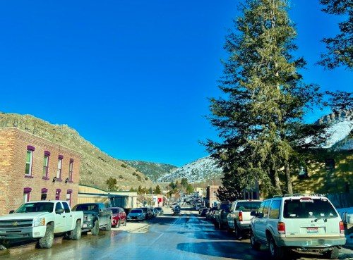 A snowy street scene with parked cars, brick buildings, and mountains under a clear blue sky.