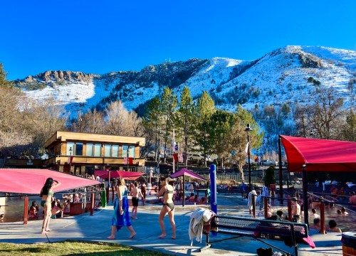 A sunny outdoor hot spring scene with people relaxing, surrounded by snow-capped mountains and trees.