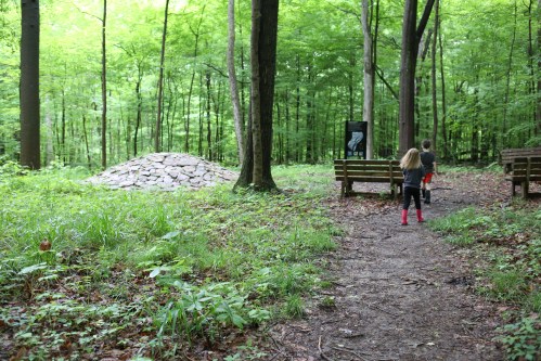 Two children walk along a path in a green forest, approaching a stone mound and benches.