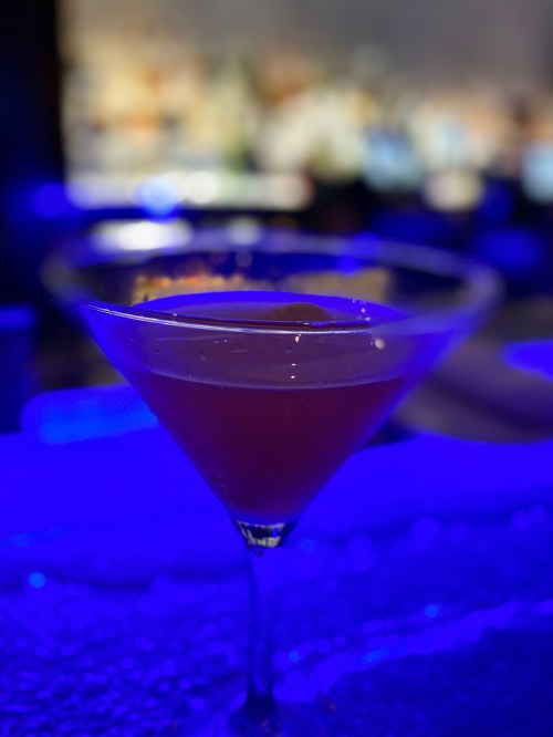A close-up of a cocktail in a martini glass, with a blurred blue-lit bar in the background.