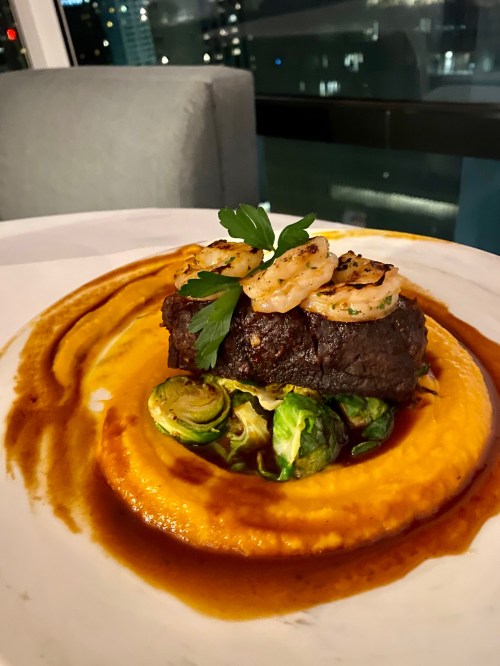 A gourmet dish featuring grilled shrimp atop a tender steak, served with Brussels sprouts and a creamy sauce.