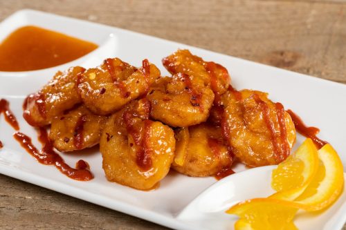 A plate of crispy shrimp drizzled with sauce, garnished with orange slices and served with a side of dipping sauce.