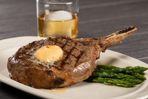 A grilled ribeye steak topped with butter, served with asparagus and a glass of whiskey on the rocks in the background.