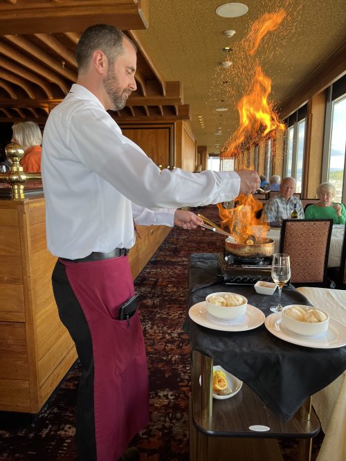 A chef in a white shirt and red apron flambés a dish at a restaurant table, with diners in the background.