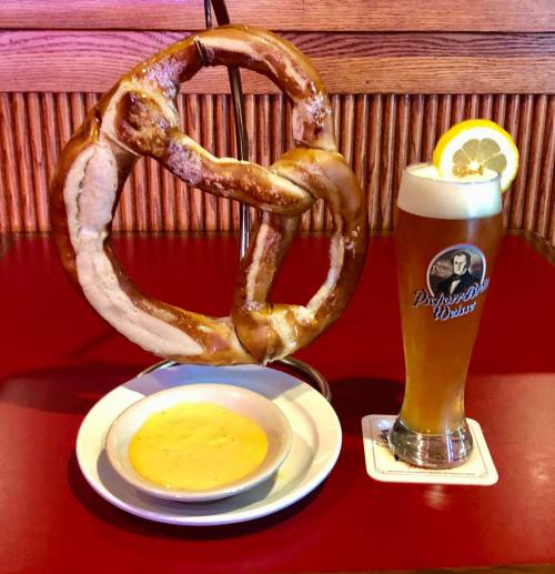 A large pretzel on a stand, served with cheese dip, alongside a tall glass of beer with a lemon slice.