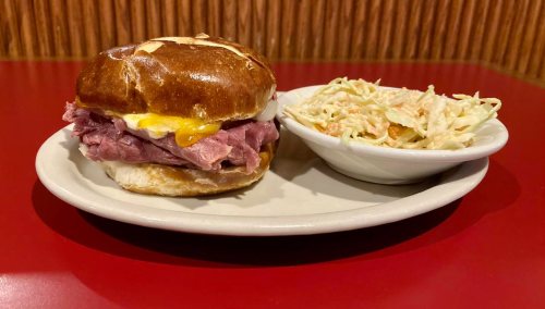 A pretzel bun sandwich filled with roast beef and cheese, served with a side of coleslaw on a red table.