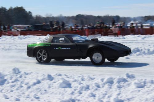 A black sports car drifts on a snowy track, surrounded by spectators and a clear blue sky.