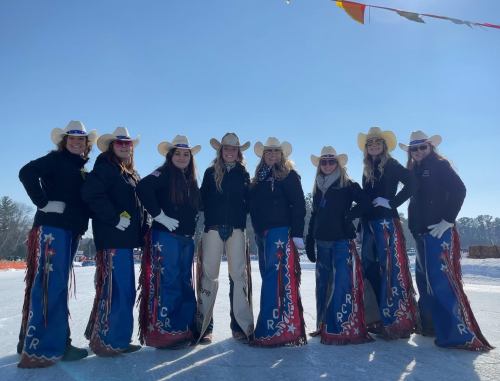 A group of eight women in cowboy hats and colorful chaps pose together outdoors on a sunny day.