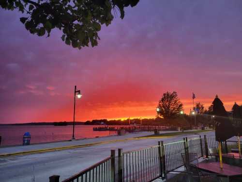 Vibrant sunset over a lake, with colorful skies and silhouettes of trees and lampposts along a waterfront path.