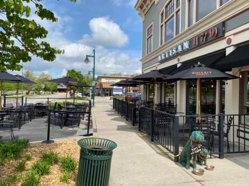 Outdoor seating area of a restaurant with umbrellas, greenery, and a decorative statue near the entrance.