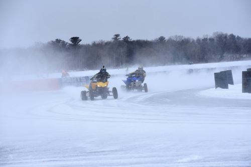 Two riders on ATVs race on a snowy track, surrounded by mist and trees in the background.