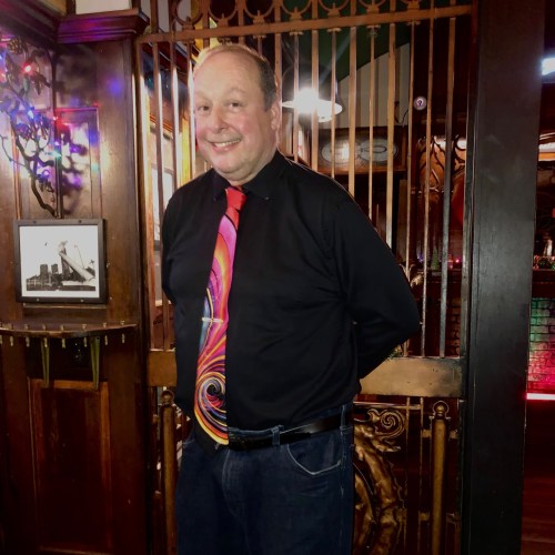 A man in a black shirt and colorful tie stands smiling in a warmly lit venue with wooden decor.