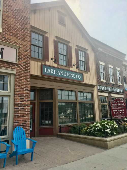 A charming storefront labeled "Lake and Pine Co." with flower beds and blue chairs outside, set in a quaint town.