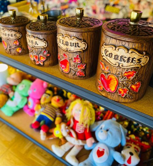 Decorative wooden canisters labeled "Coffee," "Tea," "Flour," and "Cookies," displayed on a shelf with colorful stuffed toys.